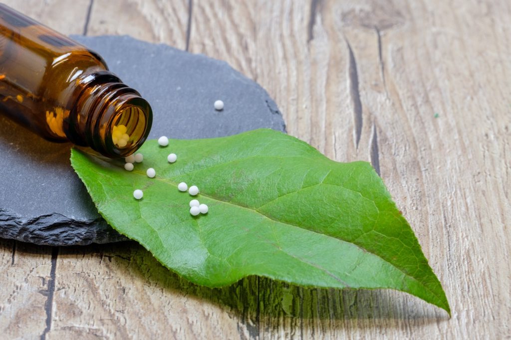 Small, white, spherical homeopathic pills spill out of their brown glass bottle, positioned horizontally on a slate, onto a green leaf