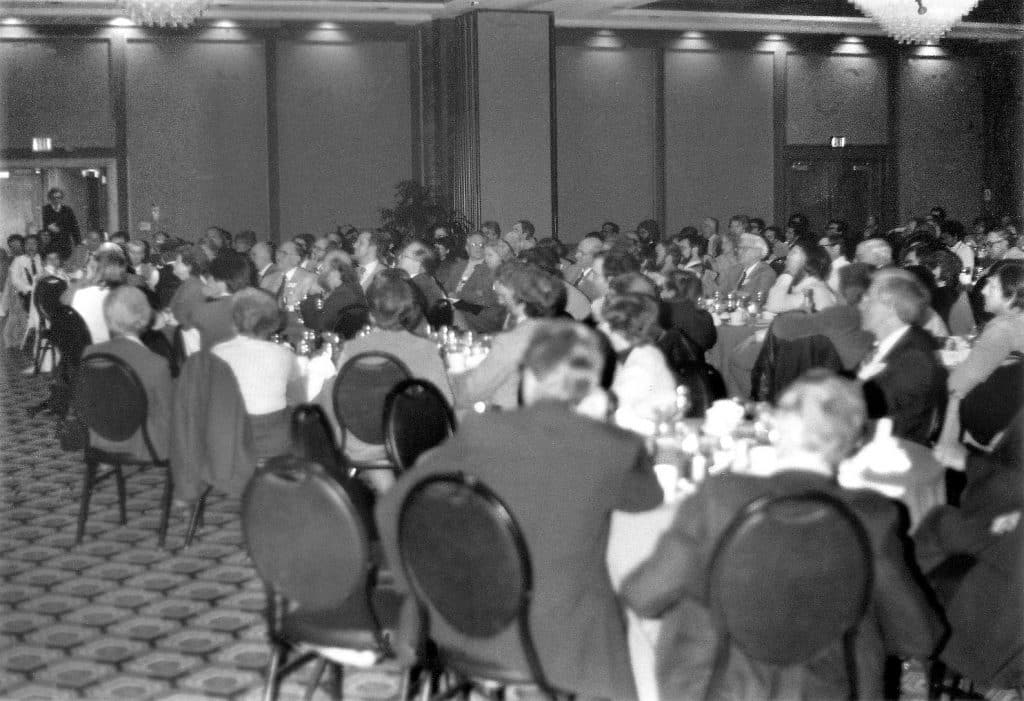 A black and white photo of a room full of people, mostly white and mostly men, sitting in a carpeted conference room on metal/leather chairs around round tables, looking towards a door as someone enters.