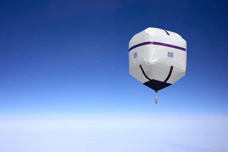 An image of a cube-shaped balloon high in the sky, with rounded corners and purple trims as well as an item hanging below it. "A visual representation of a Chinese spy balloon", by Focal Foto on Flickr (licensed under CC BY-NC 2.0 DEED)