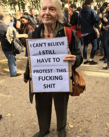 An older woman holding a sign which reads "I can't believe I still have to protest this fucking shit". 