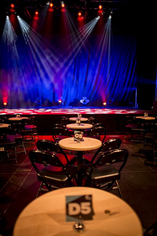 Round, wooden-top cabaret tables arranged in a line from the photographer to the stage, with more around the room. Lights shine on the stage in blue, green, red and other colours. Black folding chairs are set out behind the tables facing the stage. Labels D5, C5, B5 etc are placed on the tables.