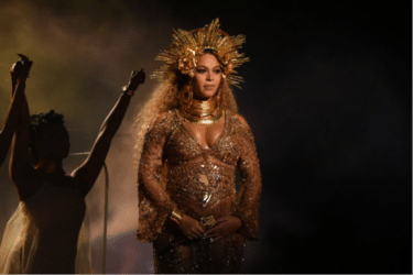 Beyonce dressed in a gold dress with a gold starburst like tiara and gold jewellery. The light is very ethereal and a woman stands in the darkness beside her with her arms raised. 