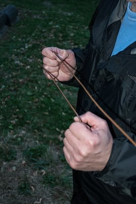 A white man is photographed standing with his hands fowards and close to his chest, grasping a dowsing rod in each hand - the rods are crossed. He's wearing a rain jacket and standing on the gradd, with a light blue sweater under his coat.