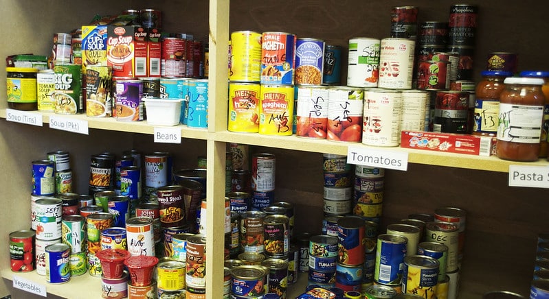 Four shelves of cans and jars of food available in a food bank, labelled as soup, beans, tomatoes, pasta sauce, vegetables etc. The donation month is written on each item in marker pen.