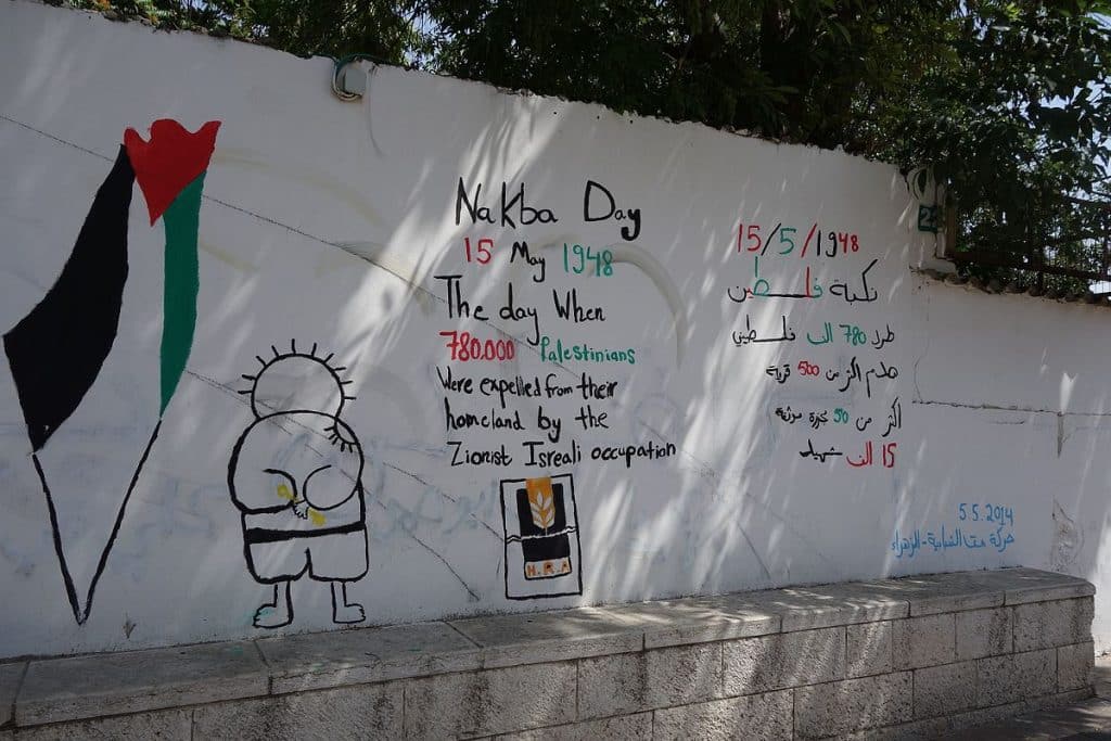 Nakba Day graffiti in black, red and green, English and Arabic, on a white wall in Nazareth, with stats/figures and a child holding a golden key behind their back, plus a stylised Palestinian flag in the shape of the region.