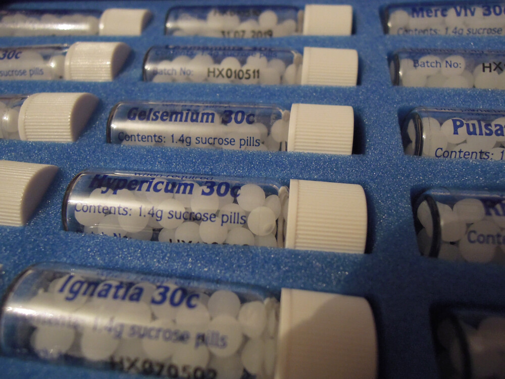 Rows of plastic homeopathic remedy vials set in blue foam, with white lids containing spherical 1.4g sucrose (sugar) pills, each labelled with the remedy name and showing 30C dilution