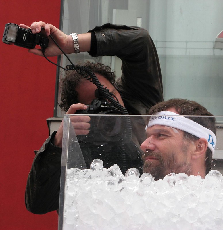 A photographer snaps pictures of Wim Hof, wearing a white headband, sat in a tank of ice that reaches up to his chin.