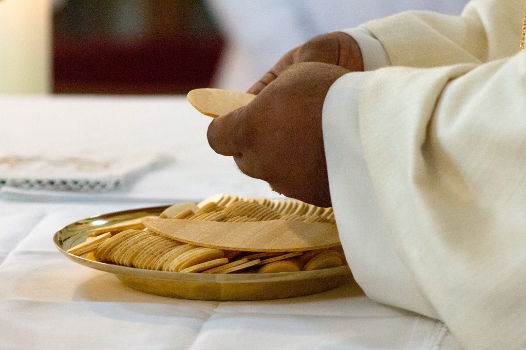 A religious official with dark-skinned, masculine hands and cream-coloured robe cuffs breaks up wafers for Eucharist