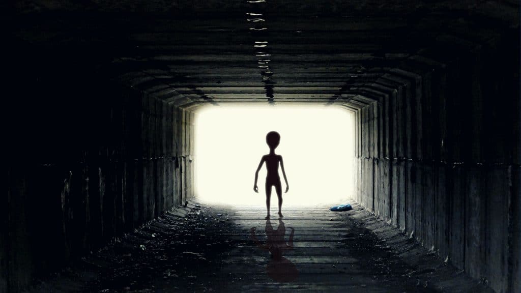 A stereotypical alien-shaped silhouette walks toward the camera through a rectangular tunnel, backlit by a bright light at its opening