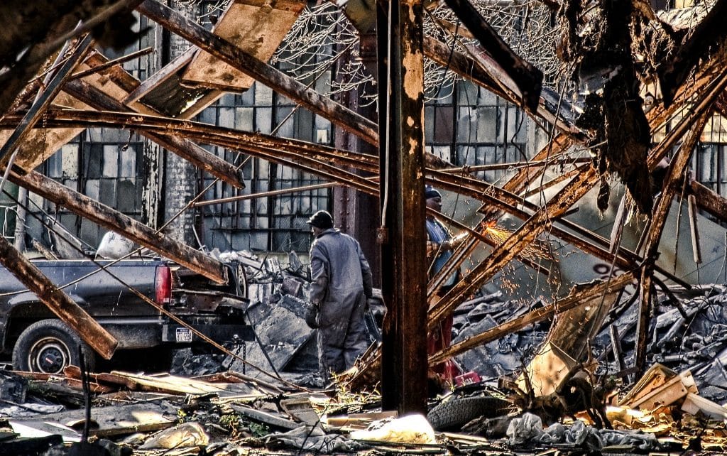 A man in overalls and gloves walks through a derelict building, patchily lit by the sun, with rusty beams and rubble all around him, back to the black pickup truck in the background. Another man to his right creates sparks as he cuts through the metal.