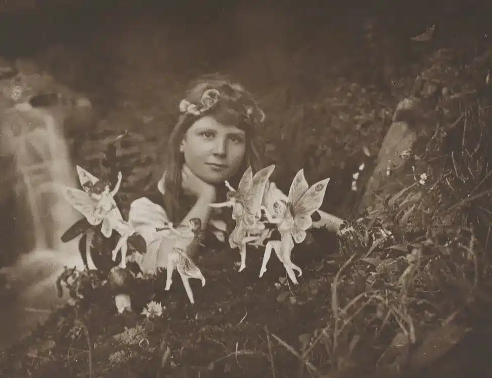 A famous 1917 sepia photograph featuring a girl with long hair decorated with some flowers, sitting in a garden in front of a waterfall, surrounded by 'fairies' (which are quite obviously not real, but many believed they were)