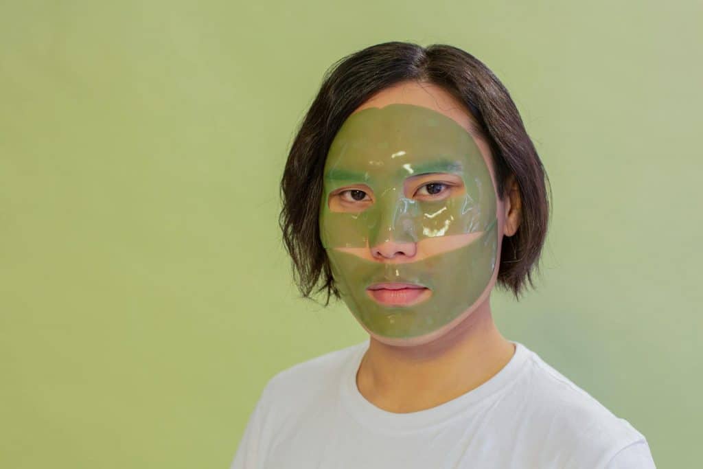 A calm-looking Asian man with jaw-length hair has a green face mask on. He's wearing a white t-shirt and standing in front of a green background