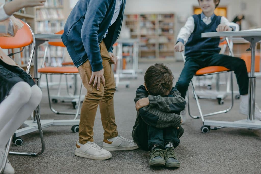 A bullied child sits on the floor with their head on their knees and arms wrapped around themseves as three other children taunt them from close by, in a classroom