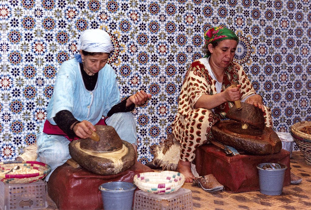 Two women sit with traditional, shaped millstones in front of them on raised platforms, grinding argan tree fruit kernels into argan paste for the production of argan oil. The tiled wall behind them has multicoloured flower-like symbols