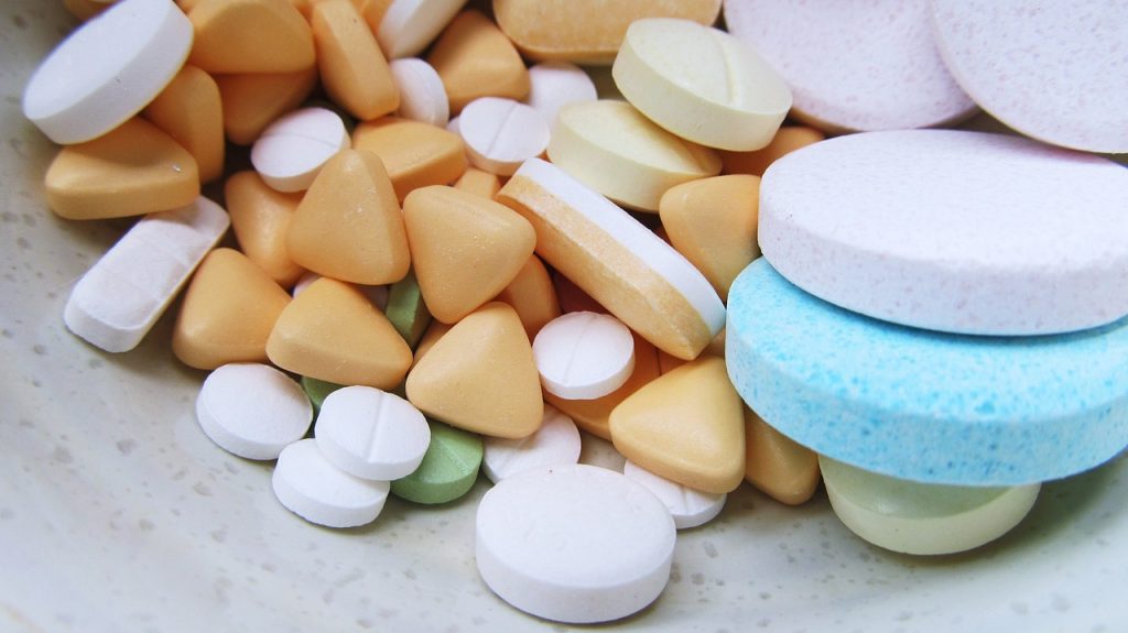 An assortment of pills in a bowl, of different shapes, sizes and colours. There are triangular, circular and capsule-shaped pills. Colours include blue, orange, green, white and pink.