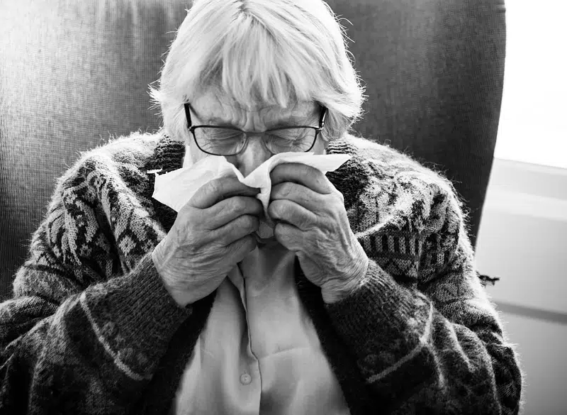 A black and white photo of a senior woman sneezing, holding a paper tissue to her face, while sitting in an arm chair wearing a cosy-looking cardigan