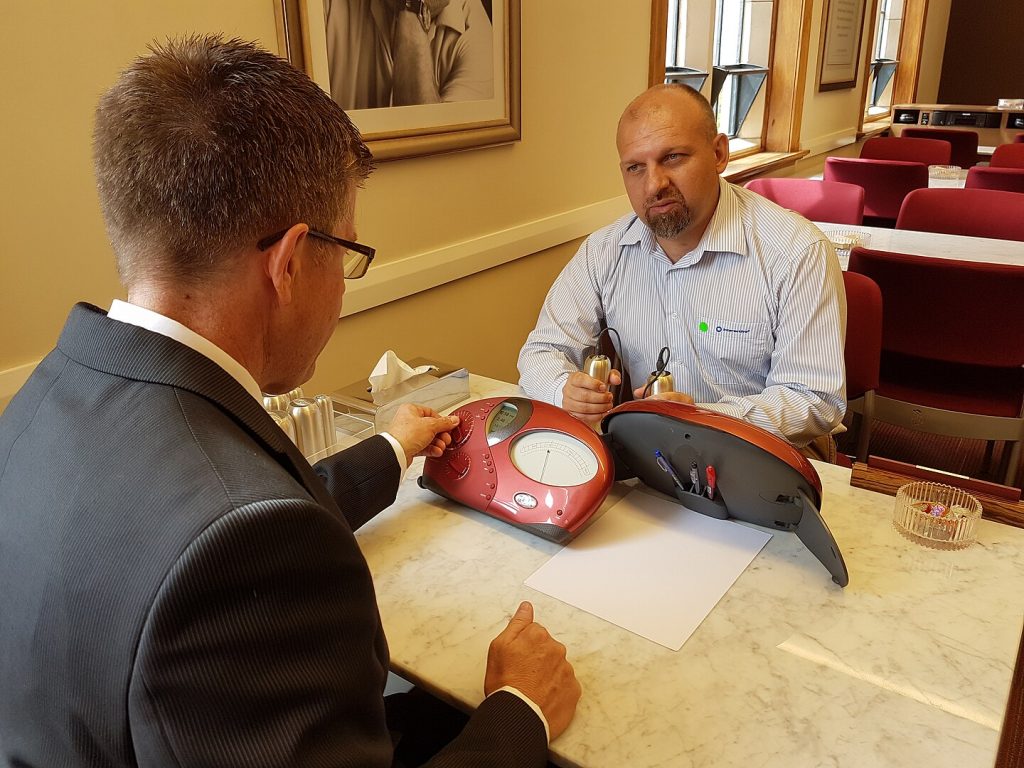 A suited Scientology auditor with glasses on sits at a table, back to the camera, adjusting a red e-meter. His subject sits across from him, holding the metallic 'cans' one in each hand, answering the auditor's questions