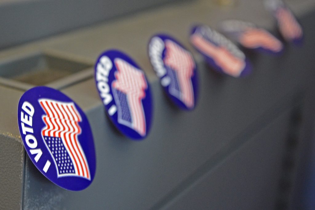 6 blue 'I VOTED' stickers with the United States of America's flag, stuck lightly to a grey metal surface. 