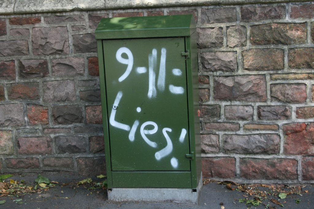 A green electrical box in front of a brick wall with white spraypaint graffiti reading "9-11: Lies!"