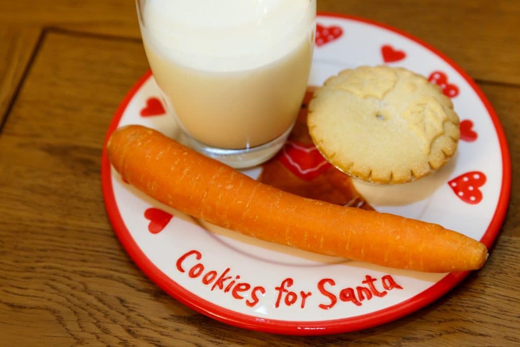 A 'Cookies for Santa' hand-decorated plate on a wooden table, with a glass of milk, a mince pie and a carrot sitting on it