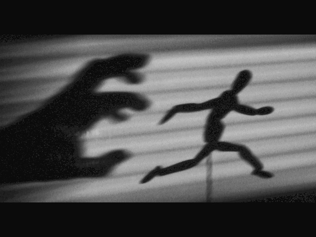 A shadow on a wall shows a menacing hand in a claw shape, reaching towards a wooden drawing aid set in a running pose. The image evokes a sense of fear and panic.