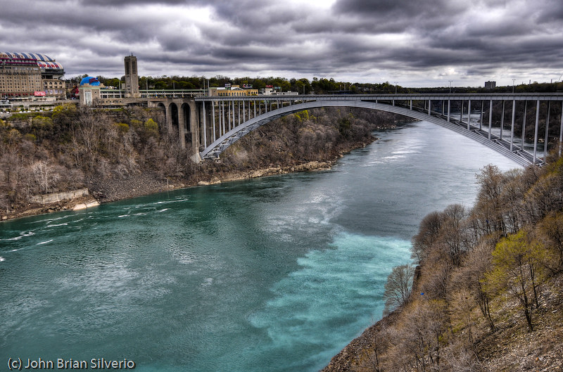 An HDR photo of the Rainbow Bridge US/Canada border crossing at Niagara, with a cloudy sky and deep teal river.