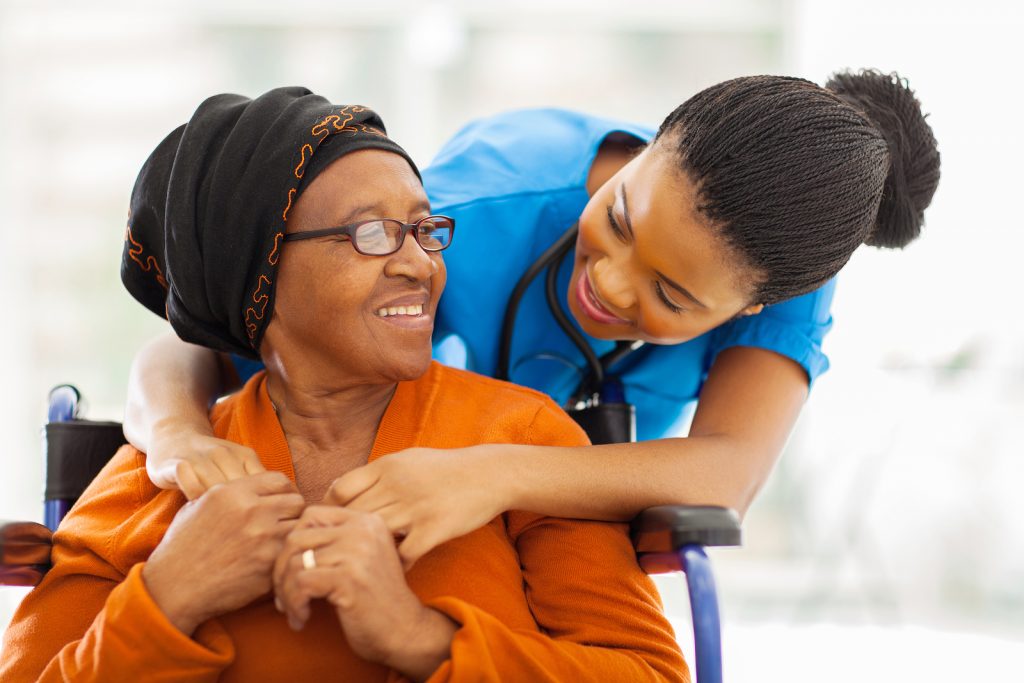 A Black female nurse in sky blue scrubs stands behind an older Black woman wearing glasses, a dark hair covering and orange top, sitting in a wheelchair, with the nurse's arms lightly wrapped around her. They hold hands, smiling at each other.