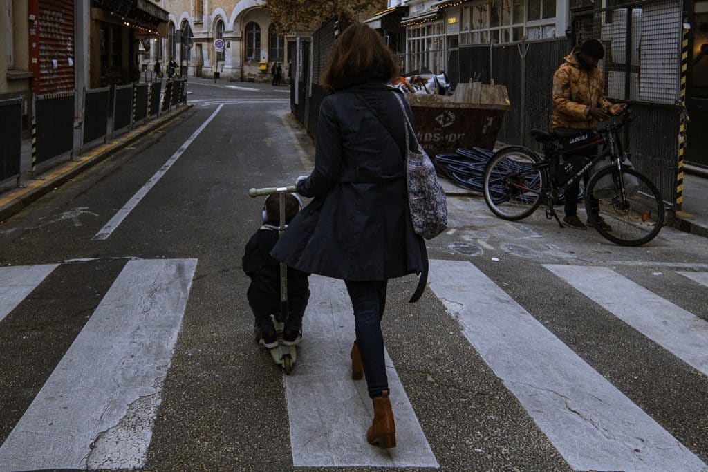 A woman guides a small child on a scooter along the centre of a Paris back street, with a man propping up his bicycle and checking his phone ahead of them, off to the side