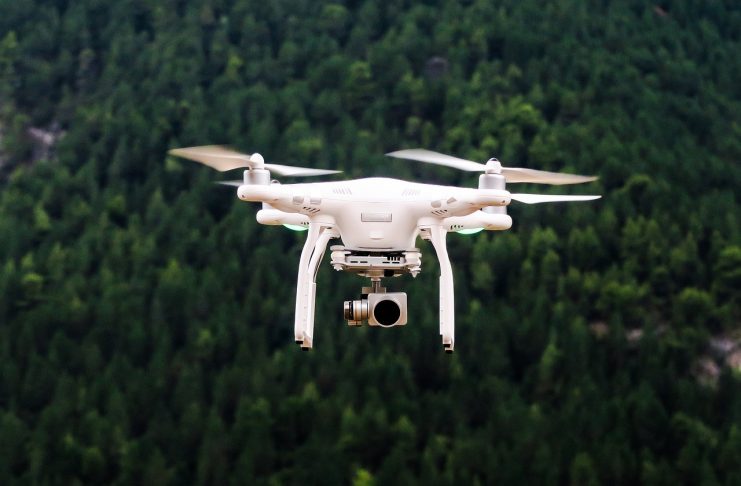 A white remote-controlled quadcopter-style drone with a camera hanging below hovers above a dark green forest