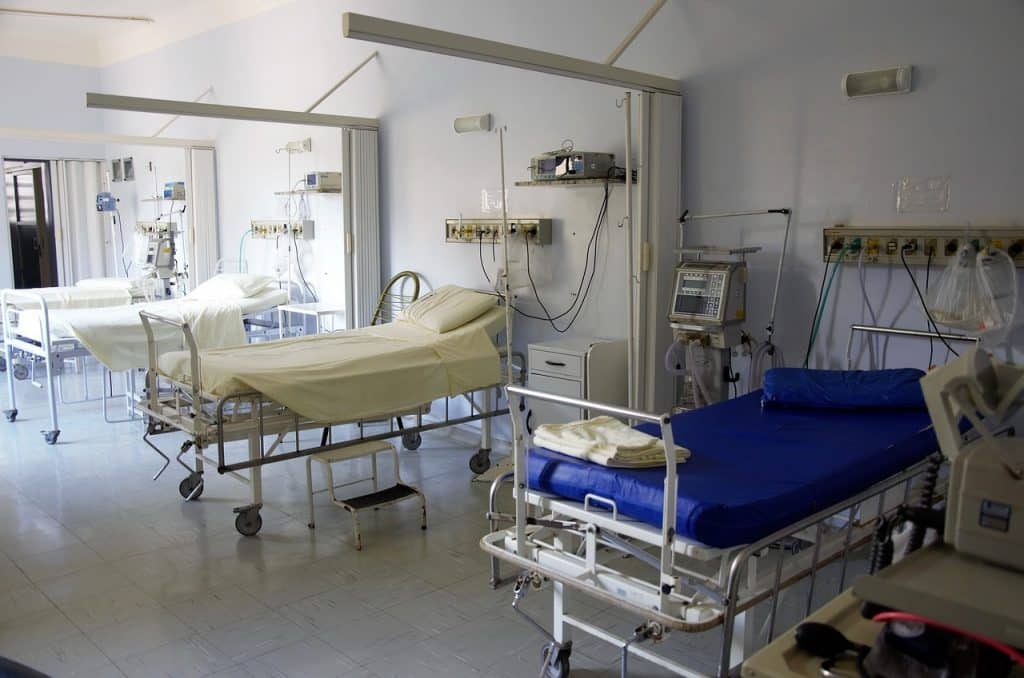 Three empty hospital beds in a ward room in daylight, two made up with bedding and one showing blue under-bedding, ready to be made.