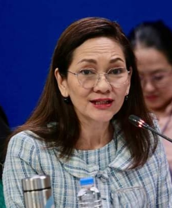 Philippines senator, Risa Hontiveros, speaks into a microphone with a thermos and water bottle in front of her. She has shoulder length, straight, dark brown hair with a side parting and is wearing colourless see-through framed glasses, red lipstick and a check shirt/dress with matching jacket