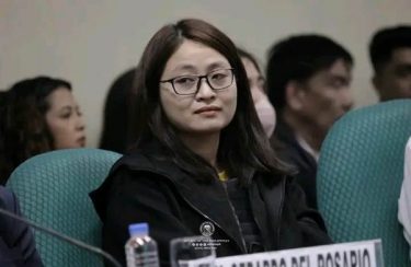 A woman, Alice Guo (mayor of a small Philippines town), sits in a green chair looking to her right, seeming unimpressed. She has straight, shoulder-length dark brown hair and is wearing square-ish framed glasses and a zip-up hoodie