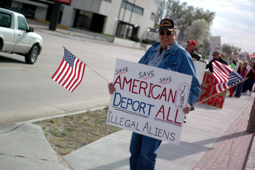 A white man in a Marine Veteran baseball cap at the 'Tax Day Tea Party in Nebraska, 2010, holds two small US flags, one in each hand, and a hand-written sign that says "SAVE SAVE SAVE AMERICAN JOBS DEPORT ALL ILLEGAL ALIENS", with all 'save's and 'deport all' underlined, and jobs off to the side encircled. More protestors are standing behind him a short way down the sidewalk.