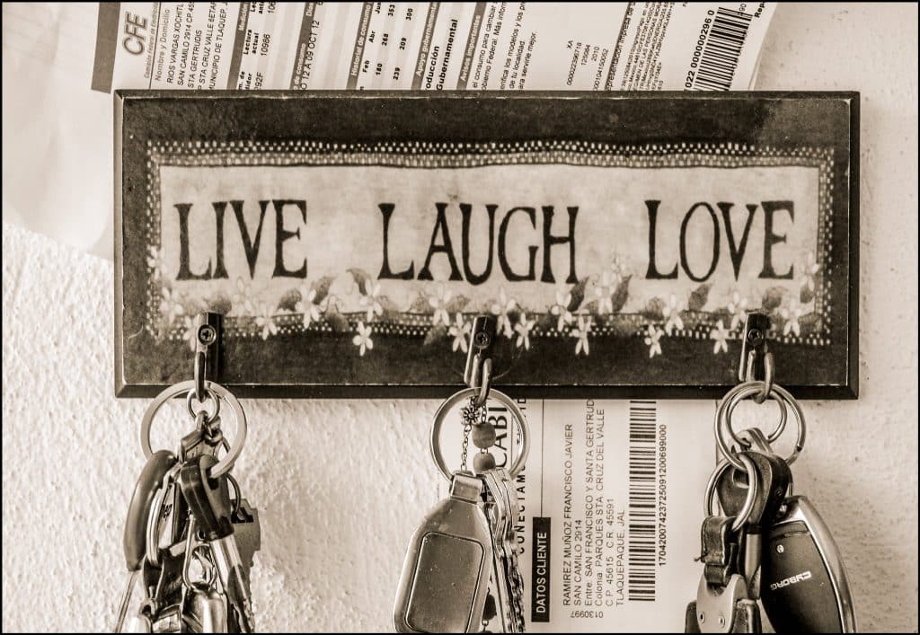 A lightly sepia-coloured monochrome photo of three keychains hanging from a 'LIVE, LAUGH, LOVE decorated plaque, which has receipts or other documents stored around it.