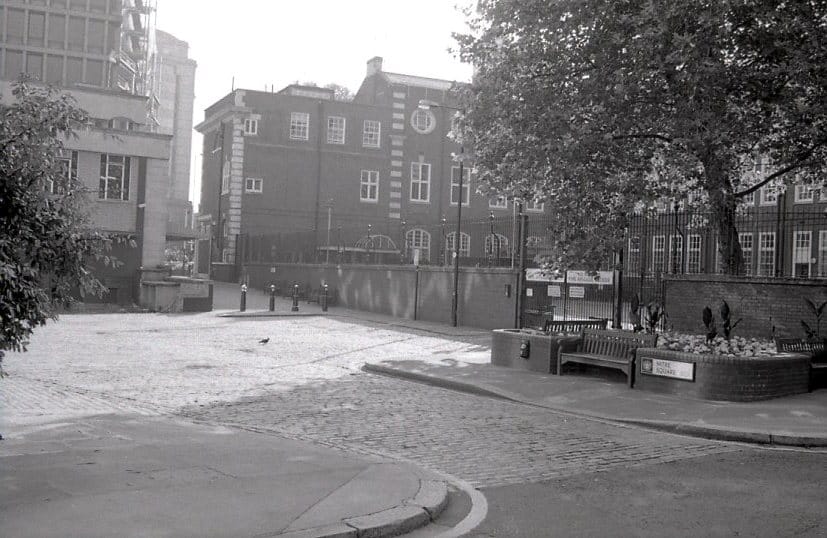 Monochrome photo of Mitre Square in London's EC3, Whitechapel. A cobbled road curves left with dark brick buildings in the background and a small park to the right. The road ahead is blocked by bollards. A road sign for Mitre Square hangs on a short wall next to a bench on the right.